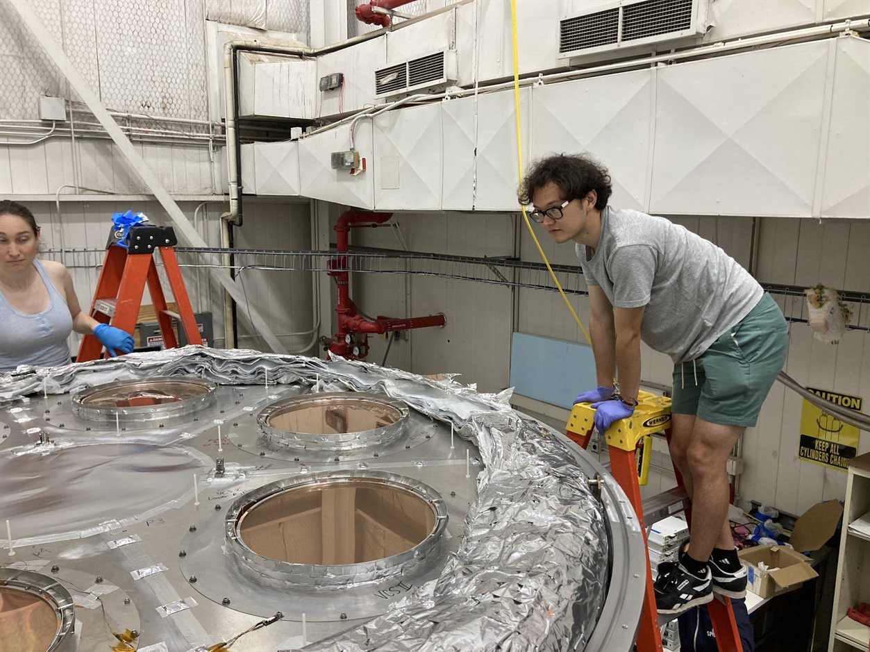 Graduate student Sho Gibbs (right) prepares to help lay and install the multi-layer insulation blanket on the uppermost layer of the cryostat. Protecting the thin plastic film filters from damage is a delicate operation.